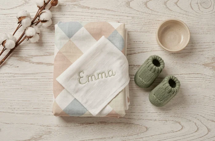 Ensemble de linge de bébé tendance : couverture swaddle en coton bio à motifs géométriques pastels, bavoir personnalisé brodé et chaussons tricotés verts sur une surface en bois blanc
